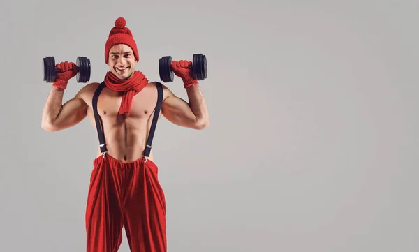 A man with a muscular figure in a red hat and a scarf with dumbbells in his hands — Stok fotoğraf