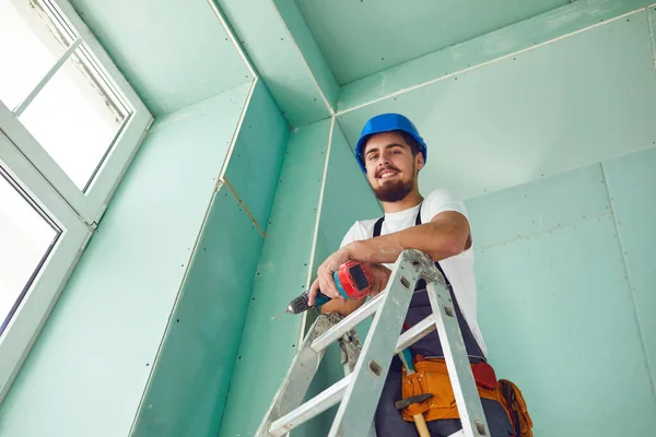 Um construtor de pé em uma escada instala drywall em um canteiro de obras — Fotografia de Stock