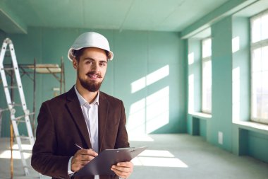 Ustabaşı sakallı adam, beyaz kasklı, inşaat alanında bir odada.