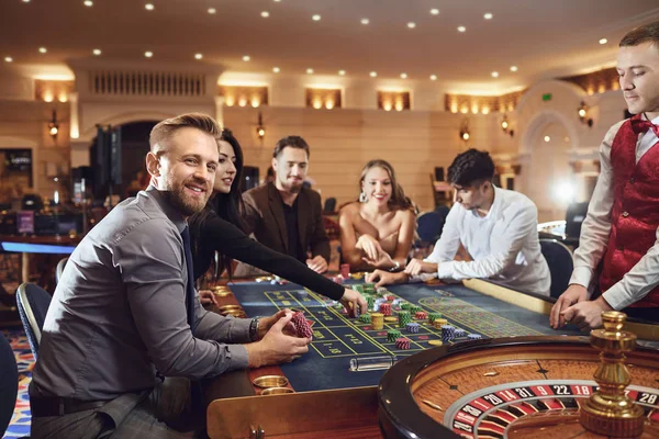 Male player smiling while sitting with friends at a poker roulette table in a casino.