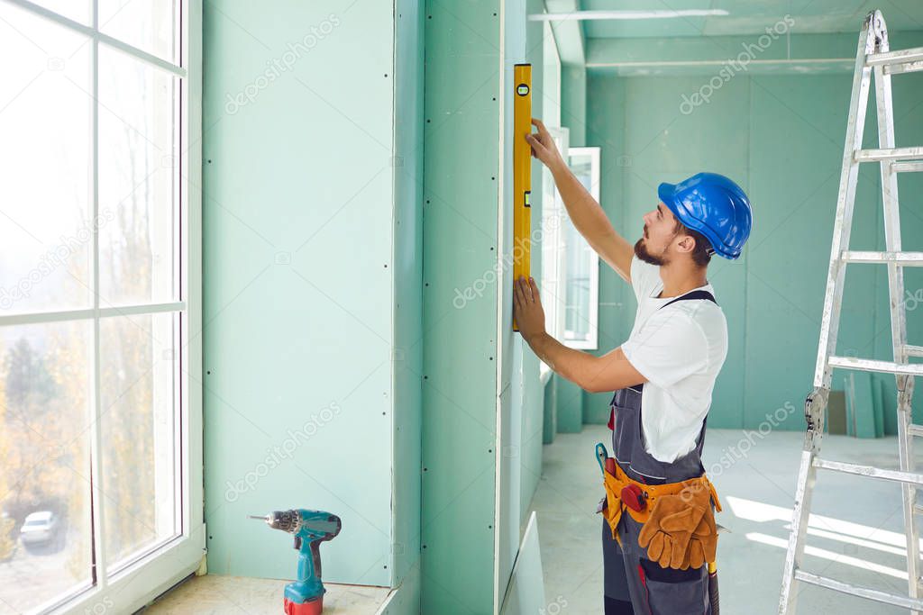 A builder standing on a ladder installs drywall at a construction site