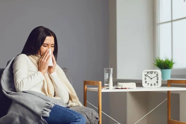 Nemocná dívka kýchající nos sedí na pohovce pod přikrývkou s příznaky chřipky virus má pilulky v místnosti — Stock fotografie