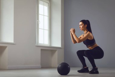 Çömelme egzersizleri. Elinde dambıllarla bir odada çömelme yapan siyah spor giysili bir kız..