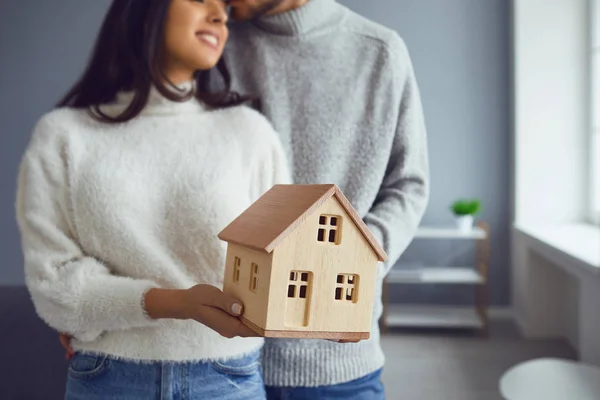 Szczęśliwa para trzymająca w rękach makietę domu stojąc w domu. — Zdjęcie stockowe