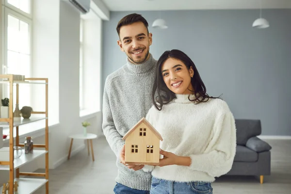 Glückliches Paar hält eine Häuserattrappe in den Händen, während es zu Hause in einem Raum steht. — Stockfoto