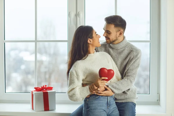 Valentines day. Beautiful smiling couple gives a heart on a background of a window in a room — 스톡 사진