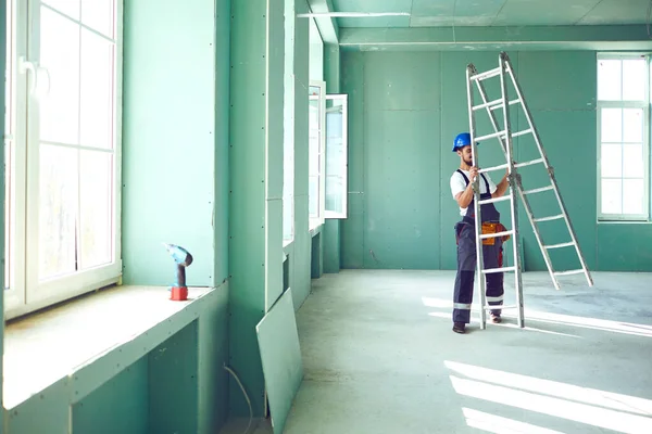 Un constructor de pie en una escalera instala paneles de yeso en un sitio de construcción — Foto de Stock