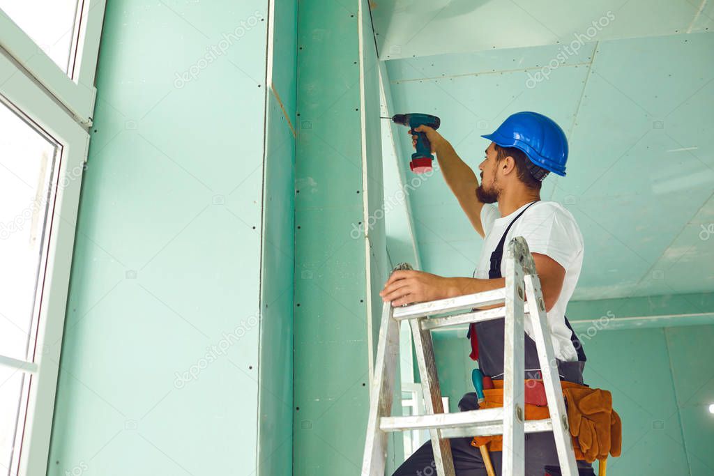 A builder standing on a ladder installs drywall at a construction site