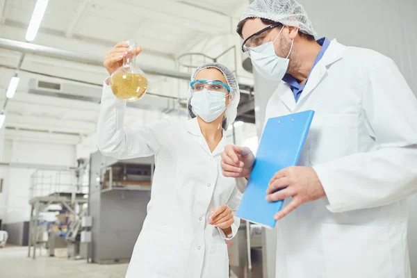 Werknemers in maskers en jassen kijken naar de vloeistof in de kolf op het werk — Stockfoto