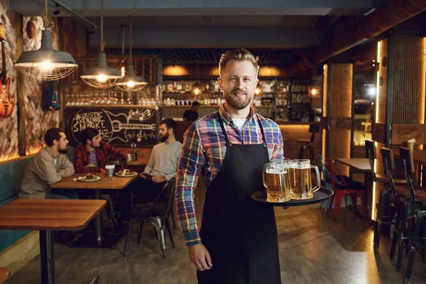 Bärtiger Kellner mit einem Tablett voller Biergläser vor dem Hintergrund einer Kneipe — Stockfoto