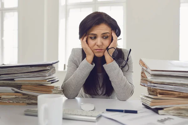 Femme brune femme d'affaires dans des lunettes avec une montagne de documents sur la table fatigué déprimé émacié déçu déprimé à la table dans le bureau — Photo