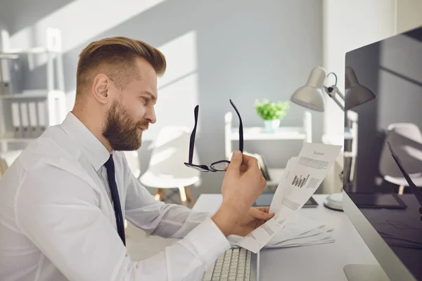 Un homme d'affaires sérieux et occupé lit des documents de travail assis à une table avec un ordinateur dans le bureau . — Photo