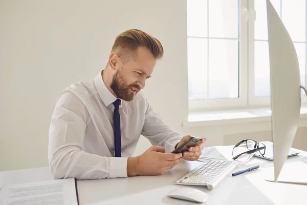 Vážný zaneprázdněný podnikatel čte zprávu na telefonu u stolu s počítačem v kanceláři. — Stock fotografie