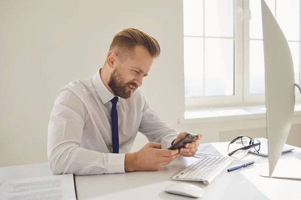 Empresário ocupado sério lê mensagem no telefone em uma mesa com um computador no escritório . — Fotografia de Stock