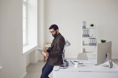 Businessman is reading in a tablet while standing at a workplace in a white office.