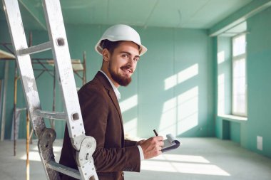 Ustabaşı sakallı adam, beyaz kasklı, inşaat alanında bir odada.