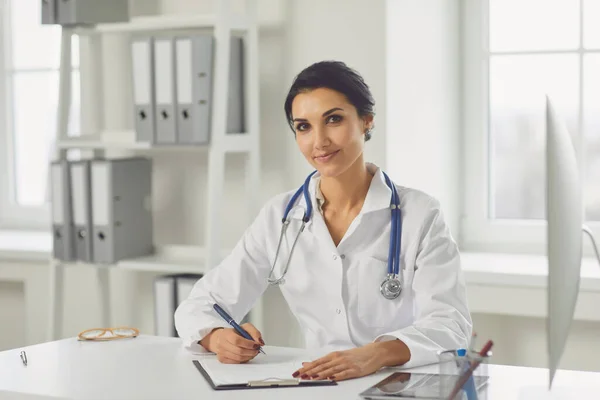 Confident woman doctor pediatrician writes in a clipboard sitting at a table in a white office of the hospital — Stok fotoğraf