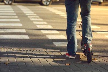 Şehrin caddesinde yaya geçidinde, elektrikli scooter 'da kot ve spor ayakkabı giyen bir adamın bacakları.
