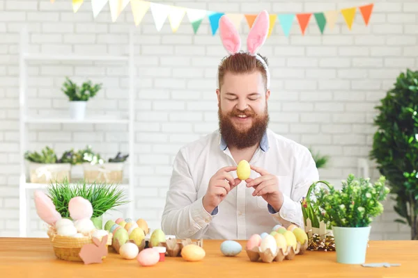 Um homem gordo engraçado decora ovos enquanto está sentado em uma mesa com decoração de Páscoa no fundo — Fotografia de Stock