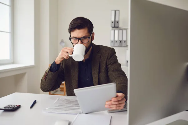 Businessman worker drinking a coffee mug while sitting at a table with a computer working in the office.