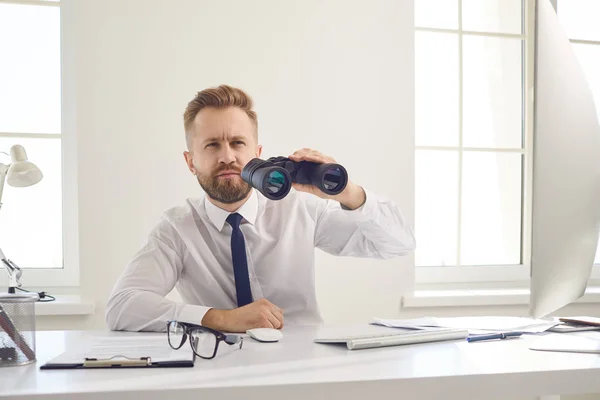 A businessman looks through binoculars while sitting at a table with a computer in the office. — Stock Photo, Image