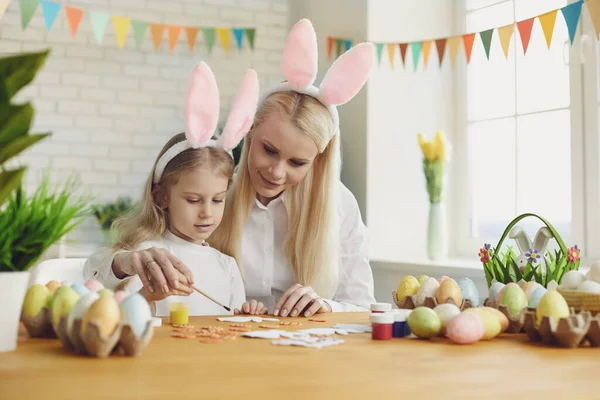 Feliz Páscoa. Filha e mãe com orelhas de coelho decoram ovos de Páscoa que se sentam em uma mesa — Fotografia de Stock