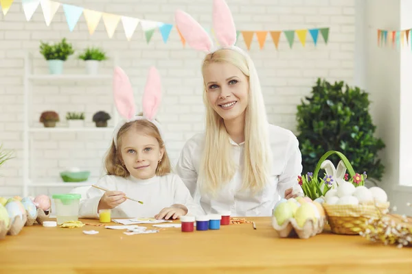 Feliz Pascua. Hija y madre con orejas de conejo decoran huevos de Pascua sentados en una mesa —  Fotos de Stock