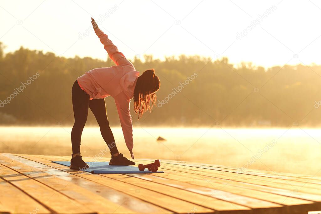 A young girl in training does squats at sunrise by the lake in autumn.