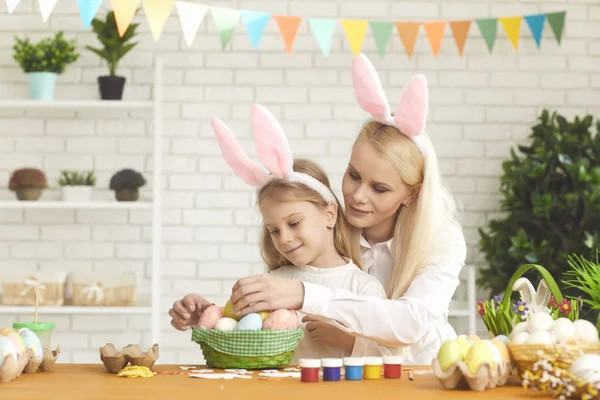 Feliz Pascua. Hija y madre con orejas de conejo decoran huevos de Pascua sentados en una mesa —  Fotos de Stock