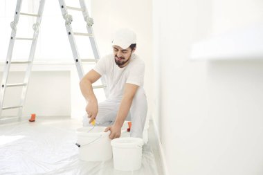 Beyaz üniformalı bir erkek ressam elinde bigudiyle beyaz bir odada çalışıyor.