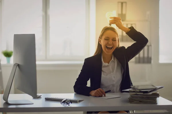 Una mujer en un traje de negocios sostiene una lámpara brillante en sus manos en una mesa en la oficina . — Foto de Stock