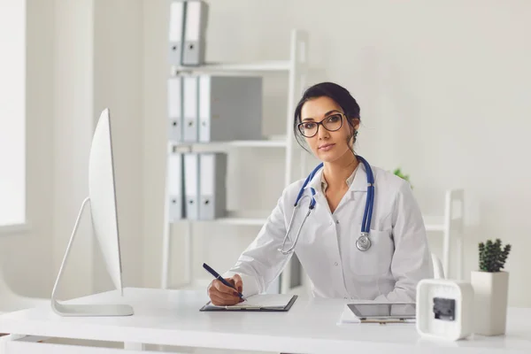 Confident woman doctor pediatrician writes in a clipboard sitting at a table in a white office of the hospital — 스톡 사진