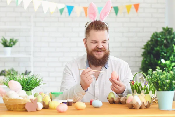 Heureux Pâques. Un homme gras drôle décore les œufs tout en étant assis à une table avec décoration de Pâques en arrière-plan — Photo