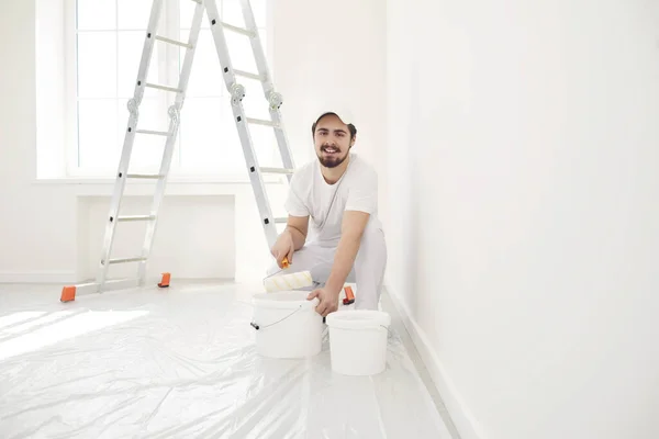 Un pintor masculino con un uniforme blanco con un rodillo trabaja en su mano en una habitación blanca — Foto de Stock