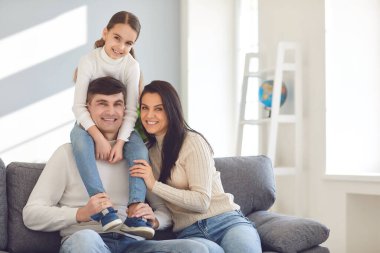 Mutlu gülümseyen aile. Kızı babasının omuzlarında, annesi de kanepede oturuyor.