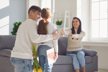 Anneler Günün kutlu olsun. Baba ve kız çocuğu, anneyi çiçeklerle ve bir odada kartpostalla kutluyor.