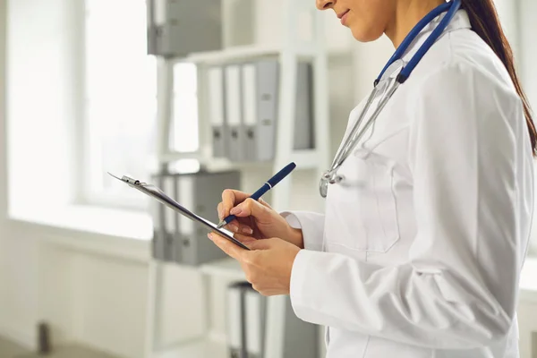 Faceless medico donna scrive una prescrizione sugli appunti mentre in piedi in ambulatorio . — Foto Stock