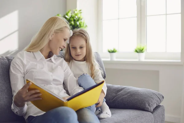 Ibu dan anak membaca buku sambil duduk di sofa di sebuah ruangan dengan jendela . — Stok Foto