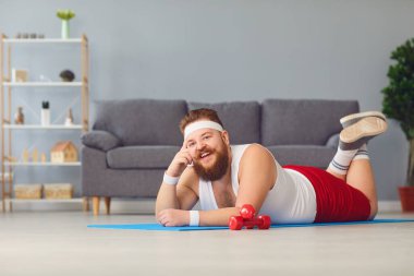 Funny red fat man doing exercises on the floor smiling while lying on the floor at home.