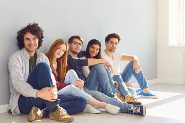 Bir grup arkadaş, gri arka planda bir odada yerde oturuyor..