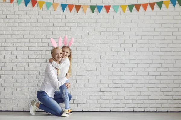 Счастливой Пасхи. Мать и дочь с кроличьими ушами, улыбающимися к белой кирпичной стене . — стоковое фото