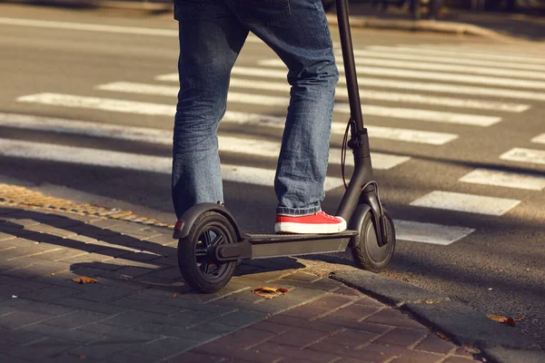 Piernas de un hombre en jeans y zapatillas en un scooter eléctrico en un paso de peatones en una calle de la ciudad —  Fotos de Stock