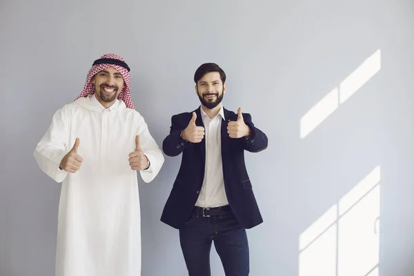 Арабские и европейские бизнесмены показывают большой палец вверх стенд улыбаясь на сером фоне . — стоковое фото
