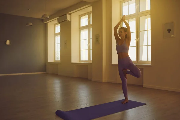 Mujer de yoga practica yoga de pie sobre una pierna medita el equilibrio en una habitación con ventanas soleadas . —  Fotos de Stock