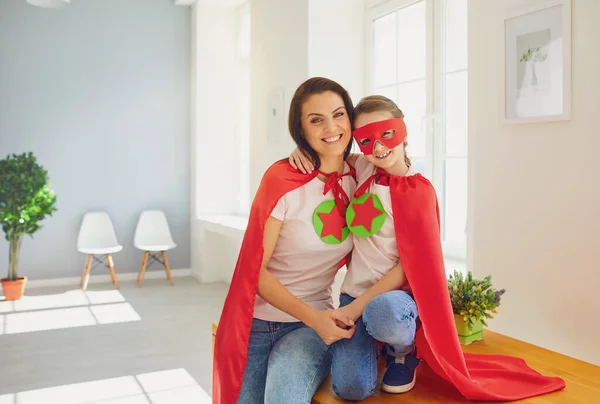 Dia das Mães. Menina e mãe em super-herói traje abraço sorrindo no quarto . — Fotografia de Stock