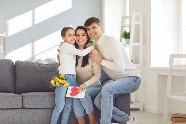 Joyeuse fête des mères. Père et bébé fille félicite maman avec des fleurs et une carte postale dans une chambre — Photo