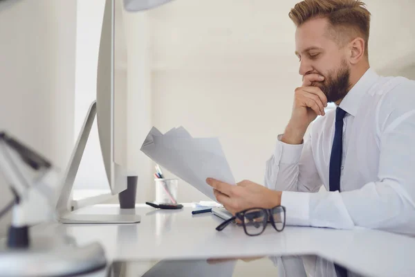 Empresário ocupado sério lê documentos de trabalho sentados em uma mesa com um computador no escritório . — Fotografia de Stock