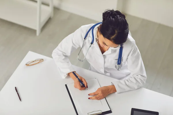 Confident woman doctor pediatrician writes in a clipboard sitting at a table in a white office of the hospital — 스톡 사진