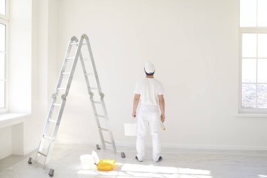 Beyaz üniformalı bir erkek ressam elinde bigudiyle beyaz bir odada çalışıyor.