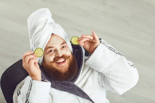 Funny fat bearded man with a cosmetic mask on his face in bathrobe towel on his head on his face resting against a gray background — 스톡 사진